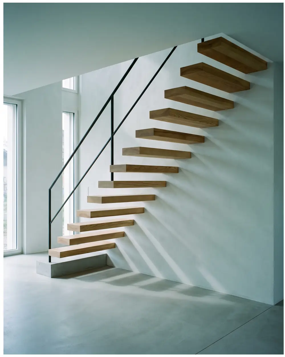Escalier suspendu en frêne massif sortant d'un mur blanc, garde-corps fin en acier noir.