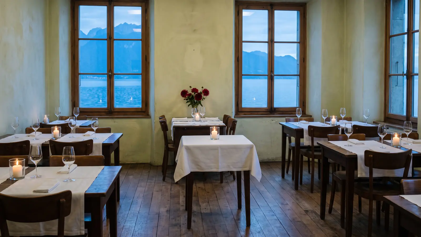 La salle de Maison Vermeil à l'heure bleue, tables dressées et bougies allumées, le lac Léman derrière les fenêtres.
